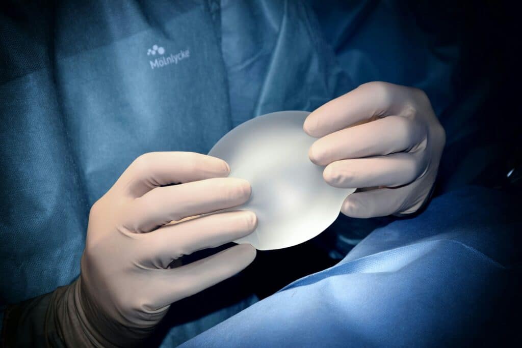 Surgeon in scrubs holding a silicone breast implant under surgical lighting, preparing for breast augmentation procedure.