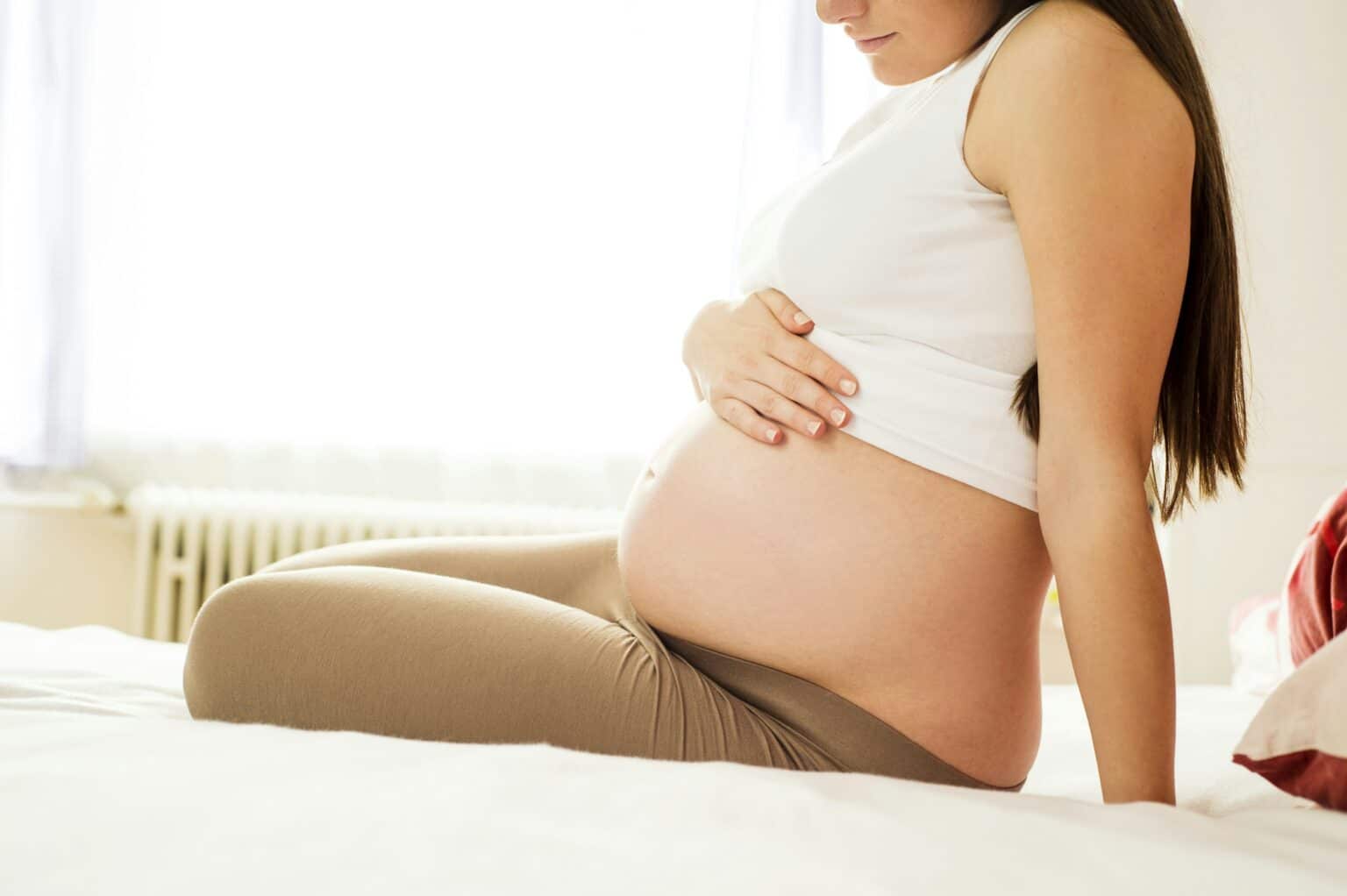 Pregnant woman sitting on a bed, gently holding her belly, symbolizing motherhood and pre-makeover transformation.