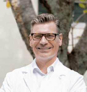 Portrait of a smiling male medical professional wearing glasses and a white coat, standing outdoors in front of a tree.