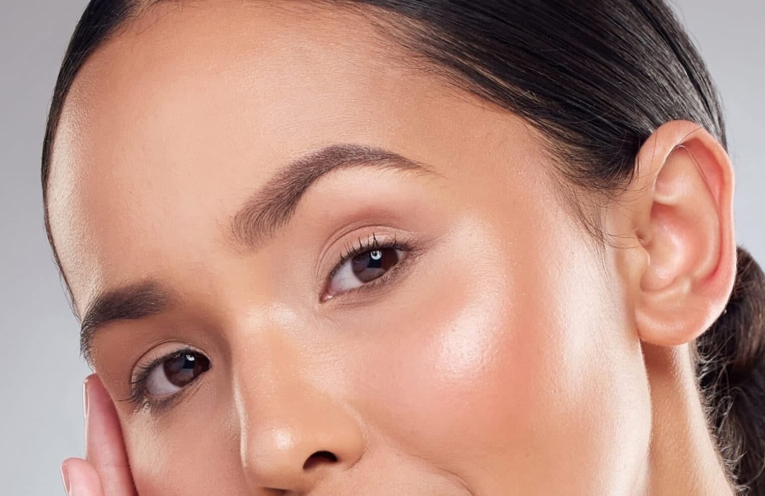 A tight close-up of a woman’s face focusing on her eyes, cheek, and ear, with dark hair pulled back smoothly. Her skin appears luminous and evenly toned with a natural glow on the cheekbone, softly groomed brows, and minimal makeup against a neutral studio background.