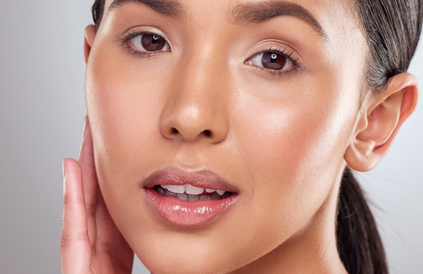 Close-up of a woman with smooth, glowing skin and dark hair pulled back, gently touching her cheek against a light gray background