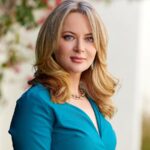 Hana Demchenko, with shoulder-length blonde hair stands outdoors, wearing a teal long-sleeve dress. She is looking toward the camera with a soft smile, with blurred greenery and flowers in the background.