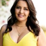 Nicolle Morazan with long wavy dark hair stands outdoors, smiling softly. She is wearing a bright yellow sleeveless dress and layered necklaces, with a blurred natural background behind her.