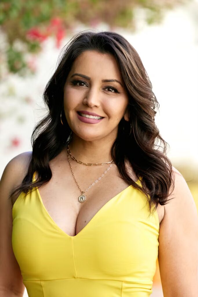 Nicolle Morazan with long wavy dark hair stands outdoors, smiling softly. She is wearing a bright yellow sleeveless dress and layered necklaces, with a blurred natural background behind her.