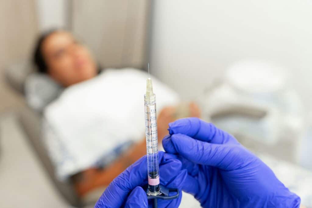 Gloved medical professional preparing an injectable treatment with a patient resting in the background.