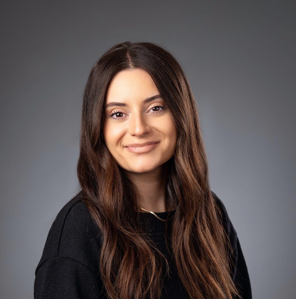 Professional studio headshot of a smiling woman with long dark brown hair, wearing a black sweater, seated against a neutral gray background with her hands folded in her lap.