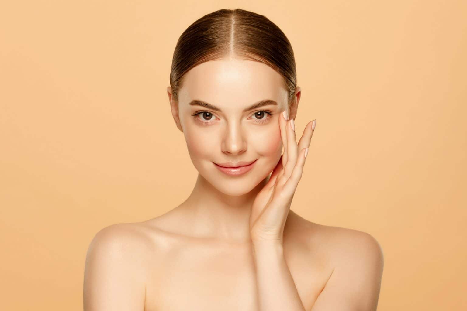 Portrait of a woman with clear, smooth skin gently touching her cheek and looking at the camera against a neutral background.